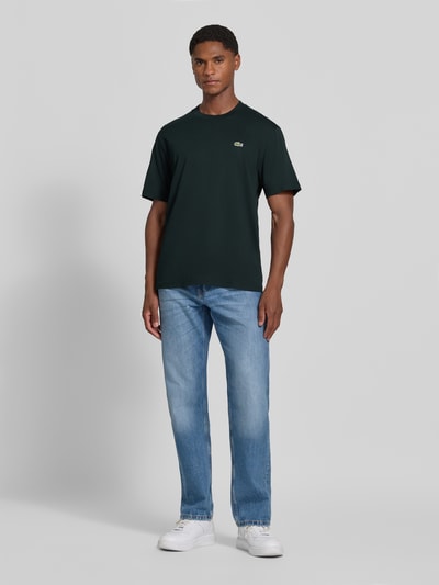 Männermodel trägt Regular Fit T-Shirt aus reiner Baumwolle von Lacoste in Dunkelgrün, kombiniert mit Jeans, Ganzkörperansicht von vorne. Herren T-Shirt von Lacoste.