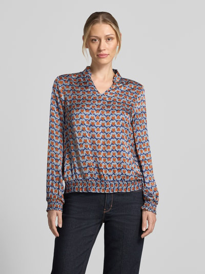 Vrouwenmodel draagt Regular Fit blouse uit zuiver viscose met geometrisch patroon van blauw, bruin en oranje van MORE, ganzichaansicht van voren. Context: dames blouse van MORE & MORE, zuiver viscose, regular fit, opstaande kraag met ruches, v-hals, met lange mouwen, elastische mouwboorden, ruglengte maat 36: 57 cm, mouwlengte maat 36: 59 cm.