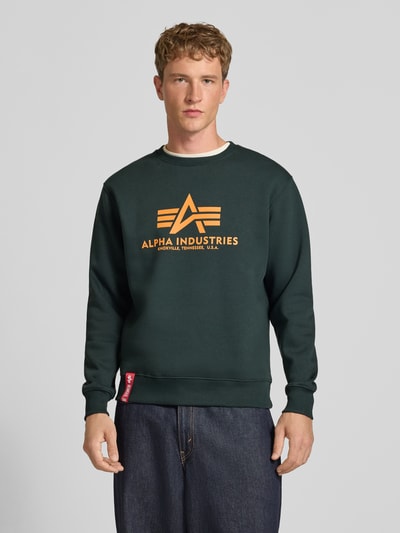 Männermodel trägt Regular Fit Sweatshirt aus Baumwoll-Mix mit Logo-Print von Alpha Industries, dunkelgrün mit orangem Print, Ganzkörperansicht von vorne.