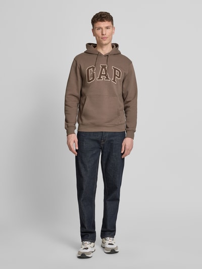 Männermodel trägt Hoodie mit Kängurutasche von GAP, braun, Baumwoll-Mix, lockere Passform, Label-Stitching, Gerippte Abschlüsse, Ganzkörperansicht von vorne.