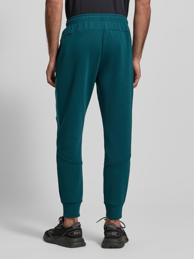 Männermodel trägt Regular Fit Sweatpants mit Logo-Print Modell 'Unstoppable' von Under Armour, Elastischer-Material-Mix, Beintasche mit Reißverschluss, Elastikbund und elastische Beinabschlüsse, Innenbeinlänge 73 cm, Farbe einfarbig dunkelgrün, Regular-Fit, Ganzkörperansicht Rückansicht.