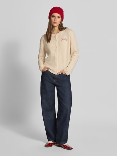Frauenmodel trägt Regular Fit Strickjacke mit Rundhalsausschnitt Modell 'EVIA' von ONLY in Beige, kombiniert mit blauer Jeans und roter Mütze, Damen Strickjacke von ONLY, Ganzkörperansicht von vorne.