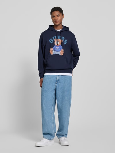 Männermodel trägt Hoodie mit Label-Print und Känguru-Tasche van GUESS in donkerblauw, katoenmix met stretch, regular fit, labelprint, kangoeroezak. Ganzkörperansicht von vorne.