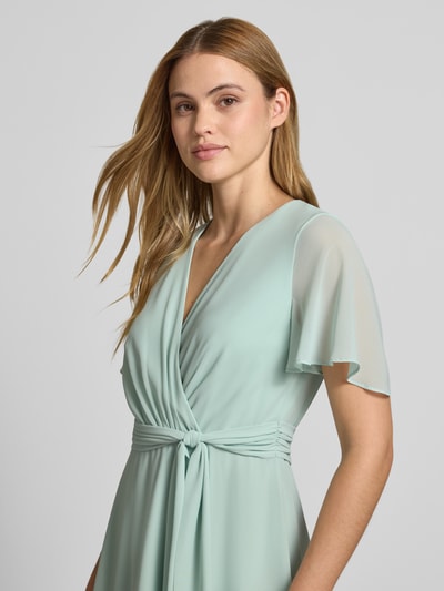 Frauenmodel trägt Knielanges Cocktailkleid in Wickel-Optik von Christian Berg Cocktail aus mintfarbenem Chiffon mit V-Ausschnitt, Bindegürtel, A-Linie, Nahaufnahme Oberkörper. Rückenlänge bei Größe 36: 100 cm, Brustweite: 33 cm, Ärmellänge: 22 cm.