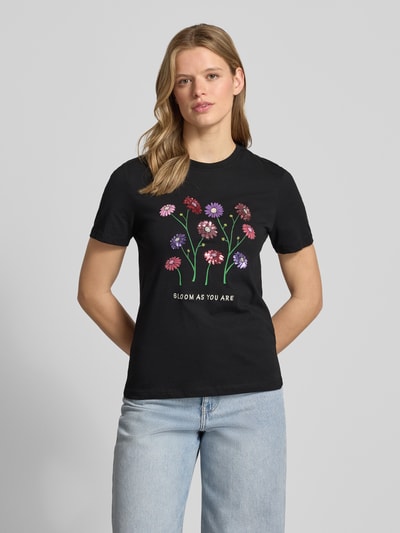 Frauenmodel trägt Slim Fit T-Shirt aus reiner Baumwolle Modell 'KITA' von Only, schwarzer Jersey, Blumen-Print, Pailletten, Ganzkörperansicht von vorne.