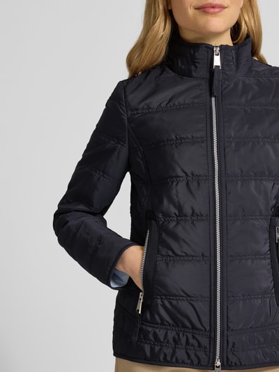 Modelka nosi czarną Steppjacke mit Stehkragen und Reißverschlusstaschen od Fuchs Schmitt, krój slim fit, dwukierunkowy zamek, boczne kieszenie na zamek, detal z logo, długość tyłu w rozmiarze 36: 65 cm, szerokość ramion w rozmiarze 36: 45 cm, szerokość w biuście w rozmiarze 36: 42 cm, długość rękawa w rozmiarze 36: 65 cm, stonowany połysk, pikowana faktura, zbliżenie na górną część ciała.