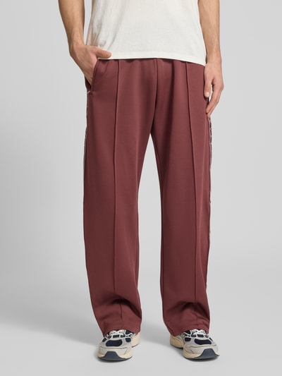 Männermodel trägt Trainingshose mit Biese, aufgesetztem Band und Logo-Bestickung von REVIEW in Rotbraun, Regular Fit, Ganzkörperansicht von vorne.