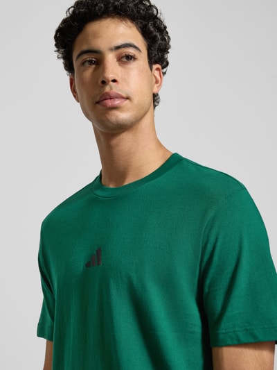 Männermodel trägt T-Shirt mit Label-Print von adidas Sportswear in Grün, reine Baumwolle, Regular-Fit, gerippter Rundhalsausschnitt, Nahaufnahme Oberkörper.