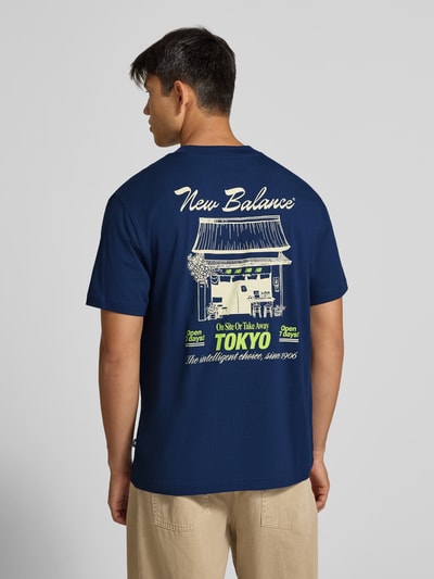 Männermodel trägt Regular Fit T-Shirt aus reiner Baumwolle mit Label-Detail von New Balance, navyblau, Rückansicht, Ganzkörperansicht. Modell 'GRAB A BITE TOKYO', Rückenlänge bei Größe M 70 cm, Print in Beige und Grün.