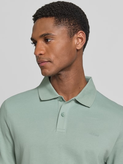 Męski model nosi Regular Fit Poloshirt aus reiner Baumwolle od s.Oliver RED LABEL, jednolity kolor, krój regular fit, detal haftu, kołnierzyk polo, dekolt w kształcie V, krótka listwa guzikowa, krótki rękaw, zakończenia rękawów i dół z bocznymi rozcięciami, tekstura pique, materiał bawełniany, zbliżenie na górną część ciała.