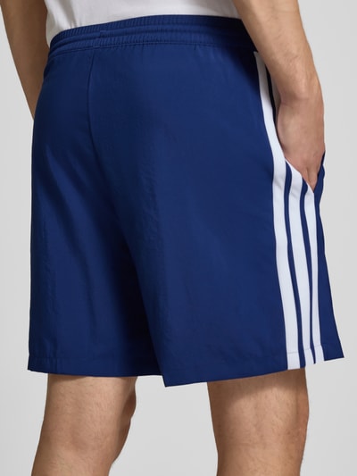 Männermodel trägt Regular Fit Sport-Shorts mit elastischem Bund von adidas Sportswear, unifarben blau mit weißen Kontraststreifen, Ganzkörperansicht Rückansicht.