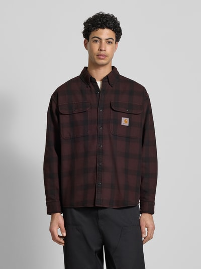 Męski model nosi Regular Fit Freizeithemd mit Logo-Patch Modell 'Edmands' von Carhartt Work In Progress, czerwono-czarna krata, bawełna, długi rękaw, kołnierzyk button-down, kieszenie na piersi, guziki, krój regular fit, widok całej sylwetki z przodu.