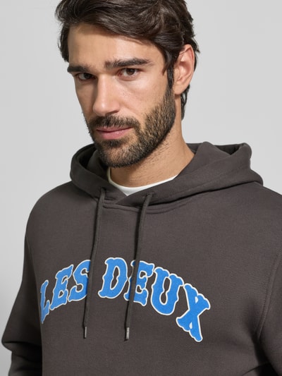 Männermodel draagt Hoodie met Label-Print model 'Brady' van Les Deux in katoen, regular fit, close-up bovenlichaam, grijs met blauwe tekst.