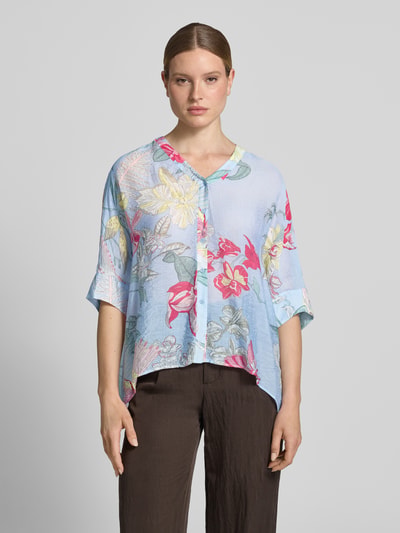 Kobiece modelka nosi Relaxed Fit Bluzę aus Viskose-Mix im Allover-Look von comma, mieszanka wiskozy, krój relaxed fit, wzór na całej powierzchni, dekolt w serek, rękaw o dł. 3/4, wydłużony tył, listwa guzikowa, długość tyłu w rozmiarze S: 64 cm, długość rękawów w rozmiarze S: 24 cm, jasnoniebieskie tło z kolorowymi kwiatami, faktura lekko strukturalna, widok całej sylwetki z przodu