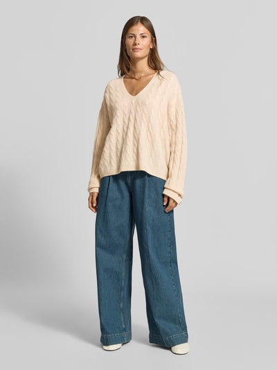 Frauenmodel trägt Relaxed Fit Pullover aus reiner Baumwolle von MYON, cremefarben, Strick-Optik, V-Ausschnitt, langärmelig, Jeans, Ganzkörperansicht von vorne.