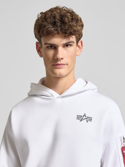 Männermodel trägt weißen Hoodie mit Label-Details von Alpha Industries aus Baumwoll-Mix, lockerer Schnitt, Nahaufnahme Oberkörper.