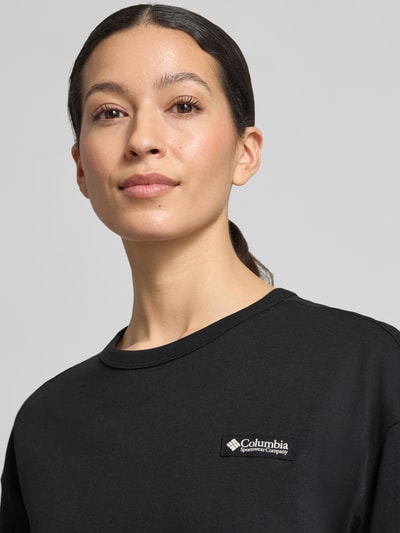 Frauenmodel trägt T-Shirt aus reiner Baumwolle mit Modell 'CSC™' von Columbia in Schwarz, Label-Print, Rundhalsausschnitt, lockere Passform, Nahaufnahme Oberkörper. Damen T-Shirt, kurzärmelig, Omni-Shade Technologie UPF 30, Rückenlänge bei Größe S 60 cm.