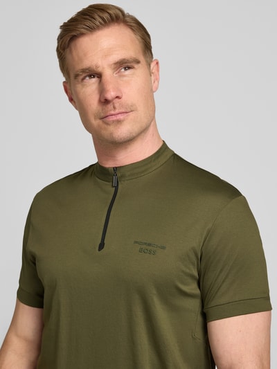 Männermodel trägt Regular Fit Poloshirt aus reiner Baumwolle Modell 'PS Spirit 70' von BOSS Green x Porsche, jednokolorowy, krój regular fit, z napisem z logo, kołnierzyk polo, zamek błyskawiczny, krótki rękaw, rozciąganie w 4 płaszczyznach, bawełna, efekt jednolity kolor, z przodu, zbliżenie na górną część ciała.