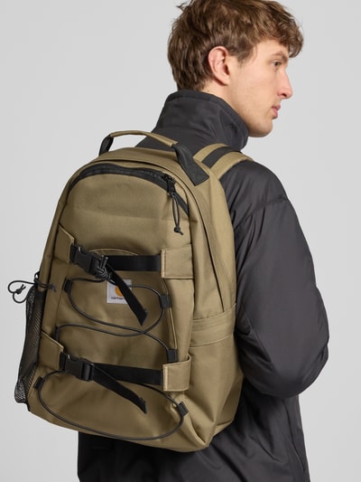 Männermodel trägt Rucksack mit Label-Detail Modell 'KICKFLIP' von Carhartt Work In Progress, oliv, Hauptfach, schwarze Schulterriemen, Ganzkörperansicht Rückansicht.