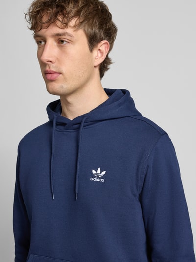 Męski model nosi Hoodie mit Kapuze od adidas Originals z bawełnianej mieszanki w kolorze granatowym, luźny krój, kaptur, tunel, label-stitching, kieszeń kangurka, ściągacze, długość tyłu w rozmiarze M: 68 cm, długość rękawa w rozmiarze M: 69 cm, zbliżenie na górną część ciała