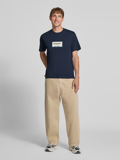 Męski model nosi t-shirt z nadrukiem z logo Model 'FREDERIKSBERG' od JACK & JONES, czysta bawełna, prosty krój, okrągły dekolt, długość tyłu w rozmiarze M: 70 cm, widok całopostaciowy z przodu.
