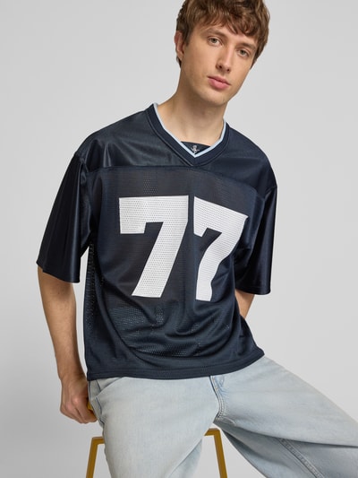 Männermodel trägt T-Shirt in semitransparenter Optik von REVIEW, dunkles Blau mit weißer 77, V-Ausschnitt, Halbarm, lockere Passform, Ganzkörperansicht von vorne.