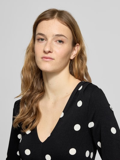 Kobiece model prezentuje Longsleeve mit V-Ausschnitt od Gina Tricot, czarny materiał w białe grochy, elastyczny materiał, dopasowany krój, rozkloszowana dolna część, dekolt w kształcie V, długość tyłu w rozmiarze S: 53 cm, zbliżenie na górną część ciała.