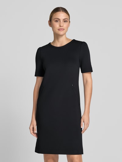 Frauenmodel trägt Knielanges Kleid mit seitlichen Eingrifftaschen von Marc Cain in Schwarz, A-Linie, Material-Mix mit Stretch-Anteil, Rundhalsausschnitt, Rückseitiger Reißverschluss, Ganzkörperansicht von vorne. Rückenlänge bei Größe 36: 87 cm, Brustweite bei Größe 36: 43 cm, Ärmellänge bei Größe 36: 26 cm.