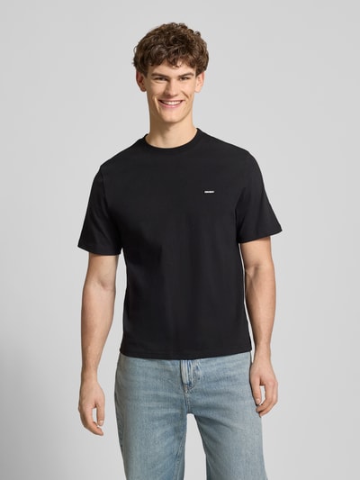 Männermodel trägt Basic T-Shirt mit kleinem Logo-Patch auf der Brust von REVIEW, schwarze Baumwolle, gerader Schnitt, Ganzkörperansicht von vorne.
