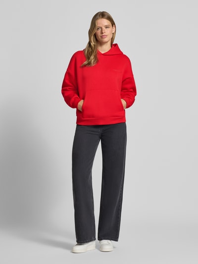 Frauenmodel trägt Kapuzenpullover aus Baumwoll-Mix von REVIEW in Rot mit Känguru-Tasche und gerippten Bündchen, Ganzkörperansicht von vorne.