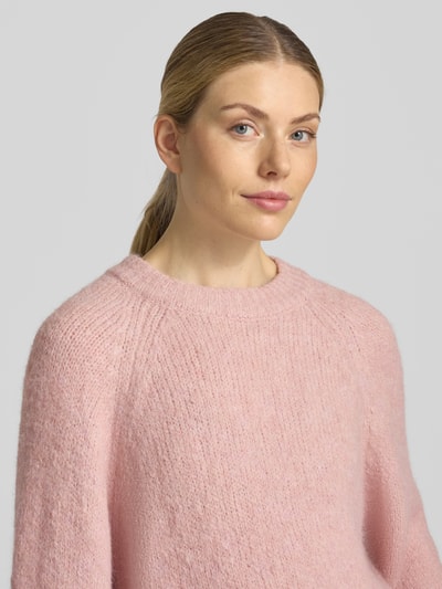 Modelka nosi sweter z dzianiny 'Strickpullover mit Raglanärmeln' od FREE/QUENT, mieszanka materiałów z dodatkiem wełny, prosty krój, okrągły dekolt, raglanowe rękawy, długość tyłu w rozmiarze S: 54 cm, długość rękawów w rozmiarze S: 50 cm, jasnoróżowy kolor, miękka tekstura, zbliżenie na górną część ciała z przodu.