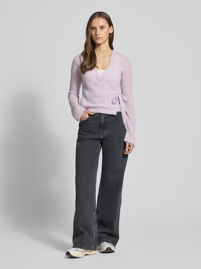Frauenmodel trägt Strick-Cardigan in Wickeloptik von Review in zartem Flieder, kombiniert mit weiter Jeans, Ganzkörperansicht von vorne. Rückenlänge 50 cm, Ärmellänge 66 cm, Schleifenverschluss, V-Ausschnitt und langer Ärmel, Regular Fit.