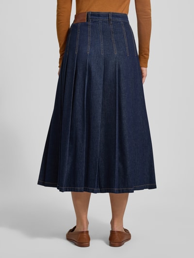 Modelka kobieca nosi rozkloszowaną spódnicę jeansową z czystej bawełny Ausgestellter Jeansrock aus reiner Baumwolle Modell 'OVALE' od Weekend Max Mara, denimowy materiał, kontrastowe szwy, długość midi, naszywka z logo, szlufki na pasek, zapięcie na guzik i kryty zamek, widoczne zakładki, widok całej sylwetki z tyłu.