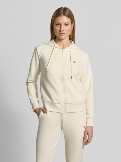 Modelka kobieca ma na sobie Sweatjacke mit Label-Applikation Modell 'OLYMPE' od Guess Activewear, kremowy kolor, luźny krój, kaptur, mieszanka materiałów z dodatkiem modalu, widoczna aplikacja logo, zdjęcie całej sylwetki z przodu.