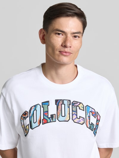 Männermodel trägt T-Shirt mit 1/2-Arm von COLUCCI aus reiner Baumwolle, weiß, bunte Buchstaben, gerader Schnitt, Nahaufnahme Oberkörper.