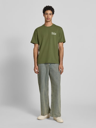 Männermodel trägt Herren T-Shirt mit Logo Modell 'VERNAL' von Dickies in Olivgrün, Reine Baumwolle, Relaxed-Fit, Rundhalsausschnitt, Ganzkörperansicht von vorne. Rückenlänge bei Größe M: 74 cm.