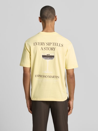 Männermodel trägt T-Shirt mit rückseitigem Motiv-Print von MCNEAL in Gelb, Rundhalsausschnitt, Statement-Print, Rückenlänge 70 cm, Ganzkörperansicht Rückansicht.