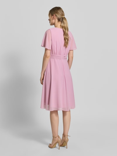 Vrouwenmodel draagt knielanges Cocktailkleid in Wickel-Optik van Christian Berg Cocktail, roze chiffon A-lijn, met bindriem, knielang, v-hals, ruglengte 100 cm, borstwijdte 33 cm, mouwlengte 22 cm, volledige lichaamsweergave achterkant.