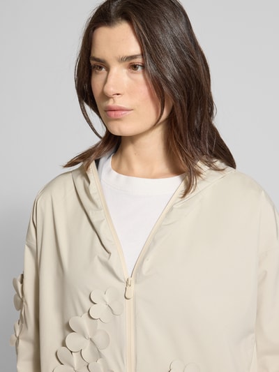 Frauenmodel trägt Regular Fit Regenjacke mit floralen Applikationen Modell 'Rosa' von SAVE THE DUCK in Beige, Logo-Patch, Reißverschluss, Nahaufnahme Oberkörper. Rückenlänge 72 cm, Schulterbreite 57 cm, Brustweite 45 cm, Ärmellänge 50 cm.