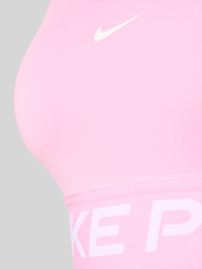 Frauenmodel trägt Sport-BH mit Label-Bund von Nike in Rosa mit weißem Logo, glatte Textur, Nahaufnahme Oberkörper, Damen Sport-BH von Nike.