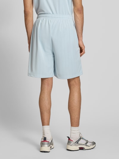 Męski model nosi Relaxed Fit Shorts z elastycznym pasem model 'Insignia' od KARL KANI, jasnoniebieskie, detal z logo, szerokość w rozmiarze M: 37 cm, długość wewnętrzna nogawki w rozmiarze M: 20 cm, widok całej sylwetki z tyłu.