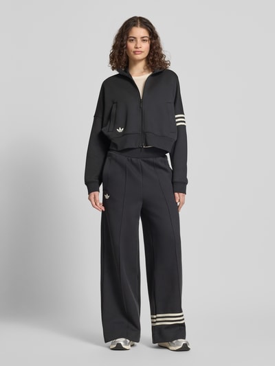 Modelka nosi Regular Fit Sweatjacke aus Baumwoll-Mix od adidas Originals, czarny, długi rękaw, suwak, logo, stójka, krój regular fit, materiał bawełniany, gładka faktura, boczne kieszenie, widok całej sylwetki z przodu.
