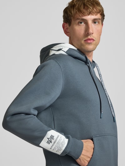 Männermodel trägt Regular Fit Hoodie aus Baumwoll-Mix mit Label-Patch von Alpha Industries, Känguru-Tasche, Nahaufnahme Oberkörper.