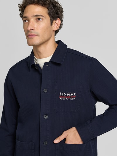 Männermodel trägt Regular Fit Hemdjacke aus reiner Baumwolle von Les Deux in Dunkelblau mit Label-Stitching und aufgesetzten Taschen, Nahaufnahme Oberkörper.