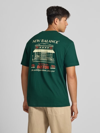 Męski model nosi t-shirt z nadrukiem z logo Model 'GRAB A BITE BOSTON' od New Balance, bawełna, prosty krój, okrągły dekolt, długość tyłu w rozmiarze M: 72 cm, widok całej sylwetki z tyłu