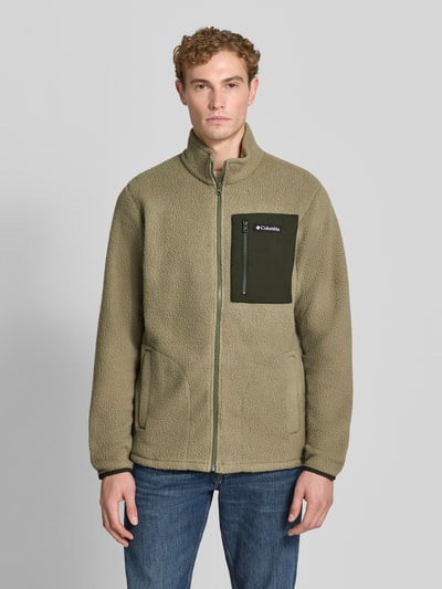 Męski model nosi kurtkę z polaru z prostym krojem, stójką, kieszenią na piersi na zamek błyskawiczny, naszywką z logo Columbia, model 'Jasper', beżowa faktura, wpuszczane kieszenie boczne, elastyczne zakończenia rękawów, długość tyłu w rozmiarze M: 72 cm, długość rękawów: 71 cm, szerokość ramion: 47 cm, szerokość w rozmiarze M: 46 cm, widok całej sylwetki z przodu.
