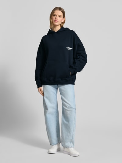 Kobieta modelka nosi bluza z kapturem Oversized Hoodie mit Label-Stitching Modell 'No Hustle' od Karo Kauer, mieszanka bawełny, krój oversized fit, kaptur, kieszeń kangurka, elastyczne wykończenia, ciemnogranatowy kolor, wyhaftowane logo, nadruk z napisem z tyłu, widok całej sylwetki z przodu.