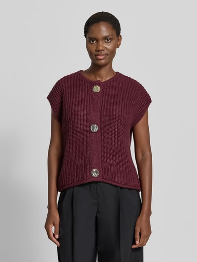 Kobiece model nosi kamizelkę dzianinową Relaxed Fit Strickweste mit Woll-Anteil Model 'Norma' od b.young, mieszanka materiałów z dodatkiem wełny, okrągły dekolt, krótkie rękawy, listwa guzikowa, długość tyłu w rozmiarze S: 52 cm, kolor bordowy, struktura grubej dzianiny, widoczne duże guziki, widok całej sylwetki z przodu.
