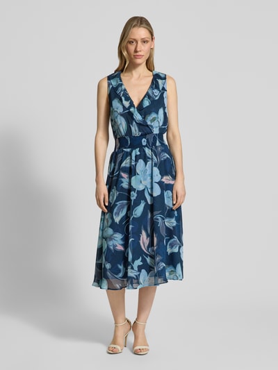 Frauenmodel trägt Midikleid mit V-Ausschnitt und Volant von More & More, taillierter Schnitt, Wickel-Optik, florales Muster, Ganzkörperansicht von vorne.