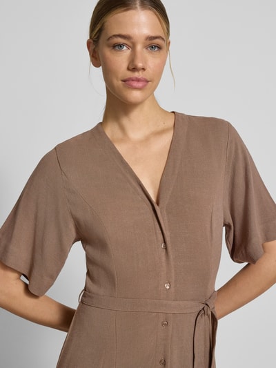 Kobiece modelka nosi Regular Fit Leinenkleid mit V-Ausschnitt Modell 'VIVA' od Selected Femme, brązowy lniano-wiskozowy materiał, gładka faktura, dekolt V, bez rękawów, pasek, guziki, kieszenie boczne, krój regular fit, maxi długość, zbliżenie na górną część ciała.